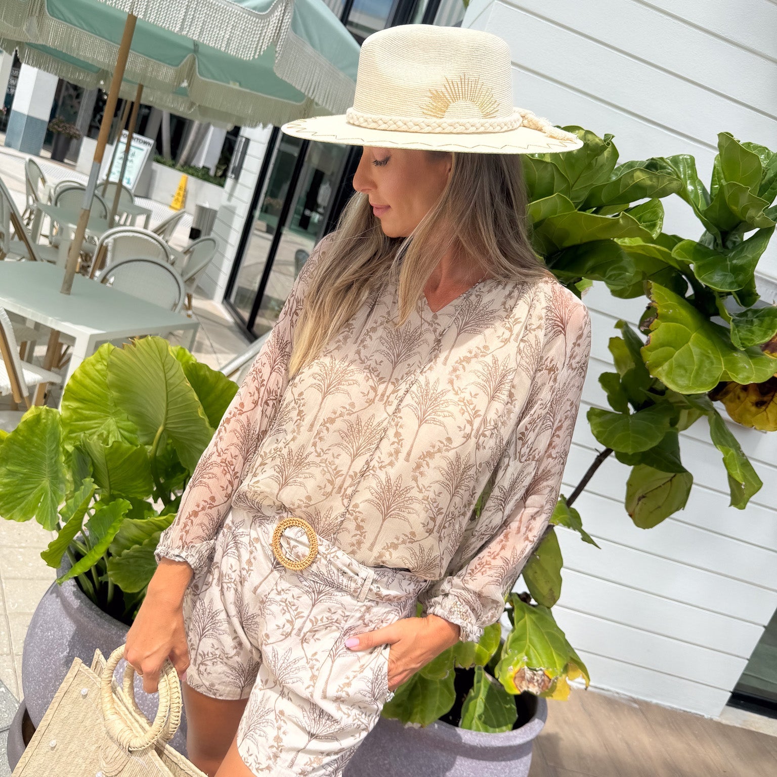 A woman wearing a beige and white patterned long-sleeve top and matching shorts with a gold-tone belt, a wide-brimmed cream sun hat, and holding a beige handbag, posing outdoors in front of a white building with green plants.